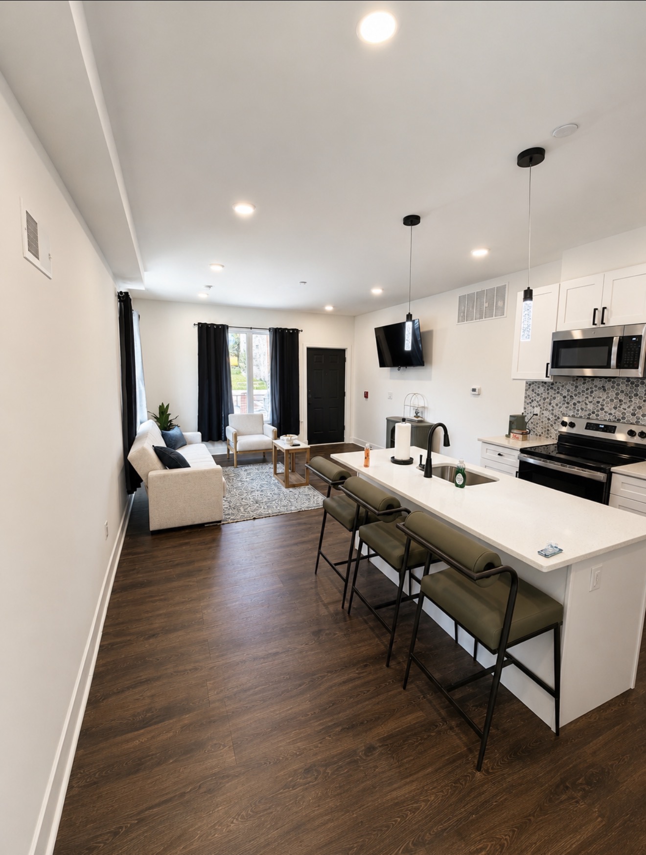Open kitchen with white island and bar stools looking into a bright living room with dark wood floors