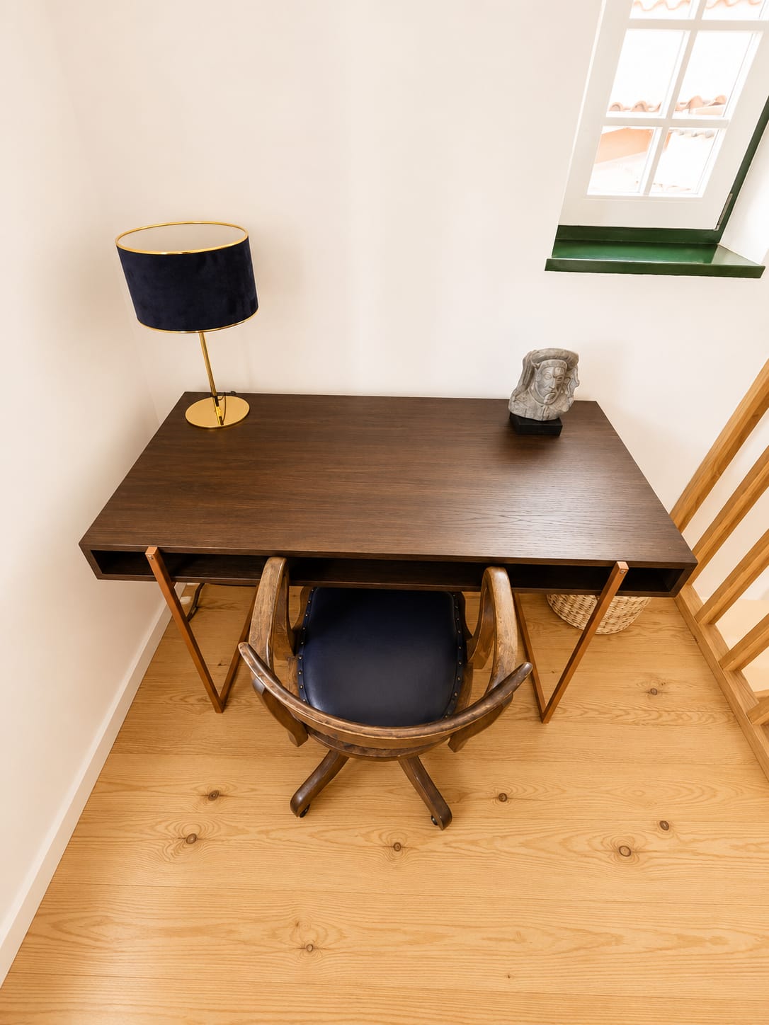 Walnut writing desk with brass lamp and swivel chair beside a green-framed window